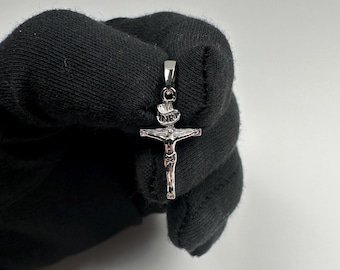 May include: A silver-colored crucifix pendant. The pendant features a detailed depiction of Jesus on the cross, with the letters "INRI" above. The pendant is held by a black glove.