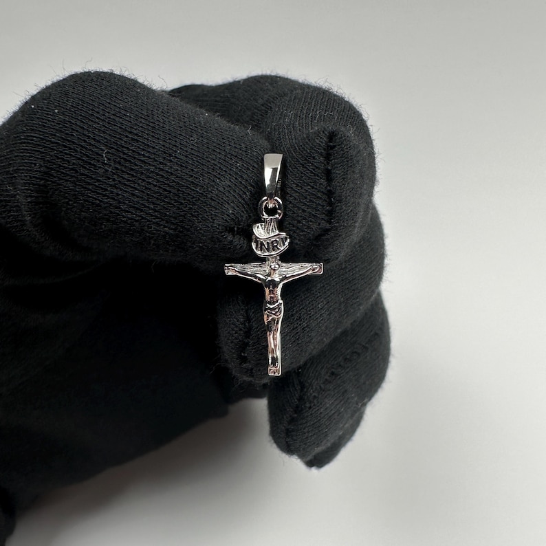 Może przedstawiać: Srebrny wisiorek z krzyżem. Wisiorek przedstawia szczeg&oacute;łowe przedstawienie Jezusa na krzyżu, z literami "INRI" powyżej. Wisiorek jest trzymany przez czarną rękawicę.