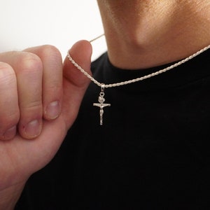 May include: Silver crucifix pendant necklace. The pendant shows Jesus on the cross. The necklace is held by a person wearing a black top. The chain is a thin, twisted rope style.