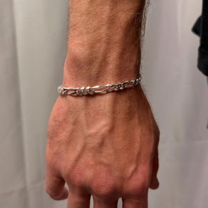 May include: A silver Figaro chain bracelet is displayed on a wrist. The bracelet features a repeating pattern of elongated and rounded links. The bracelet is made of a shiny, reflective metal.