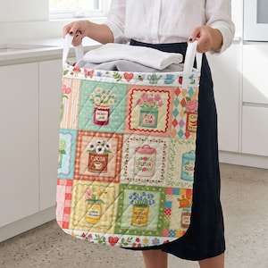May include: A quilted fabric laundry hamper featuring a patchwork design of colorful kitchen canisters and jars labeled with text including Berry Jam, Tea Tin, Cocoa, Flour, Sugar, Lemon Preserves, and Mustard Seed, with white fabric handles.