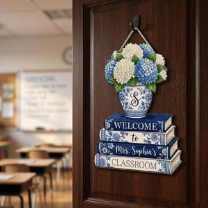 May include: A hanging wall sign featuring a stack of four books with blue and white floral patterns. The top book reads Welcome, the second to, the third Mrs. Sophia's, and the bottom CLASSROOM. A blue and white vase with hydrangeas sits on top.