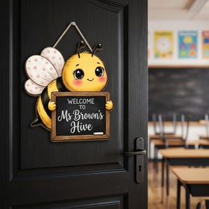May include: A decorative wall sign shaped like a cartoon bee with yellow and black stripes, pink cheeks, and white polka-dot wings. The bee holds a rectangular chalkboard with the text "WELCOME TO Ms Brown's Hive" written in white.