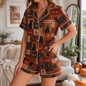 May include: A short-sleeve button-down pajama set featuring a patchwork pattern of black cats, pumpkins, moons, and stars in shades of orange, brown, and black. The shirt includes a chest pocket and white piping trim throughout the garment.