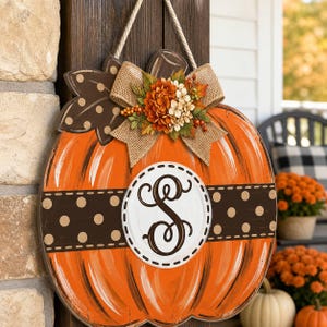 Puede incluir: Un colgador de puerta de madera con forma de calabaza en color naranja brillante con una banda horizontal marrón oscuro decorada con lunares beige. Un círculo blanco central muestra una letra S negra. Un lazo de arpillera y flores decoran la parte superior.