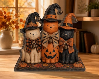 Puede incluir: Una figura decorativa de mesa con tres gatos sentados sobre una base rectangular. Los gatos llevan sombreros puntiagudos y lazos de cuadros con cascabeles. El gato central es naranja con una cara de calabaza, flanqueado por un gato crema y uno negro.