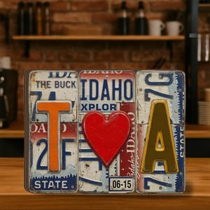 Könnte beinhalten: Ein dekoratives Stück, das "IDA" mit wiederverwendeten Nummernschildern buchstabiert. Das "T" ist orange, das Herz ist rot und das "A" ist goldfarben. Die Nummernschilder haben blaue, weiße und rote Akzente.