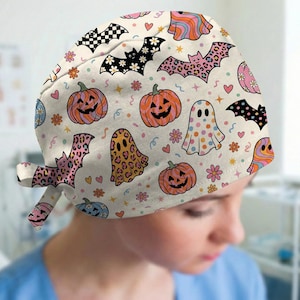 May include: A scrub cap with a cream background featuring a repeating pattern of orange jack-o'-lanterns, ghosts with colorful polka dots or leopard print, and bats with floral, checkered, or leopard patterns, accented by small pink hearts and flowers.