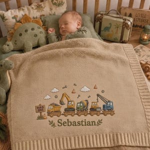 May include: A beige knit baby blanket featuring an embroidered design of construction vehicles on a train track, including a dump truck, excavator, cement mixer, and crane. The text "Build Zone" and the name "Sebastian" are embroidered in brown.