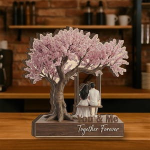 May include: A wooden sculpture of a tree with pink blossoms, a swing, and two figures. The base reads "You & Me Together Forever". The tree trunk has the names "Sophia & Henry" engraved on it. The sculpture is on a wooden surface.