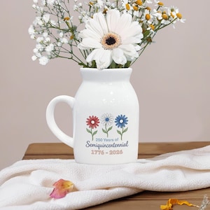 May include: A white ceramic vase with a handle, decorated with red, white, and blue flower graphics and the text "250 Years of Semiquincentennial 1776-2026." The vase holds a bouquet of white flowers.