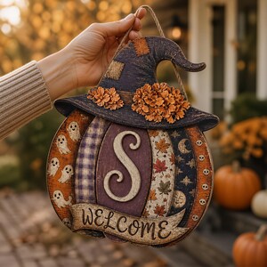 Puede incluir: Un colgante de pared con forma de calabaza y un sombrero de bruja con detalles florales naranjas. La calabaza est&aacute; dividida en paneles verticales con patrones de fantasmas, cuadros, la letra S, hojas de oto&ntilde;o, lunas y calaveras, con una pancarta que dice WELCOME.