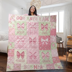 May include: A pink and green quilt with the words "GOD SAYS I AM" at the top. The quilt features bow designs and words like "WORTHY", "COURAGEOUS", and "LOVED". The name "SOPHIA" is at the bottom. The quilt is displayed by a person.