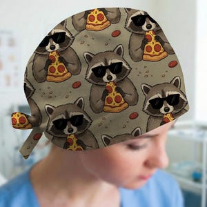 May include: A gray surgical scrub cap featuring a repeating pattern of cartoon raccoons wearing sunglasses and eating pizza slices. The design includes scattered pepperoni and a light gray background.