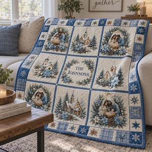 May include: A quilted throw blanket featuring a grid of nine panels with blue and white winter themes. The panels display nativity scenes, churches, and pine trees, with a central panel reading THE JOHNSONS surrounded by a blue floral wreath.
