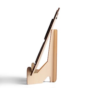 Puede incluir: Un soporte de exhibici&oacute;n de madera de color claro visto desde un perfil lateral. El soporte cuenta con un respaldo vertical y una repisa frontal dise&ntilde;ada para sostener un objeto plano en un &aacute;ngulo inclinado. La construcci&oacute;n consiste en paneles de madera entrelazados.