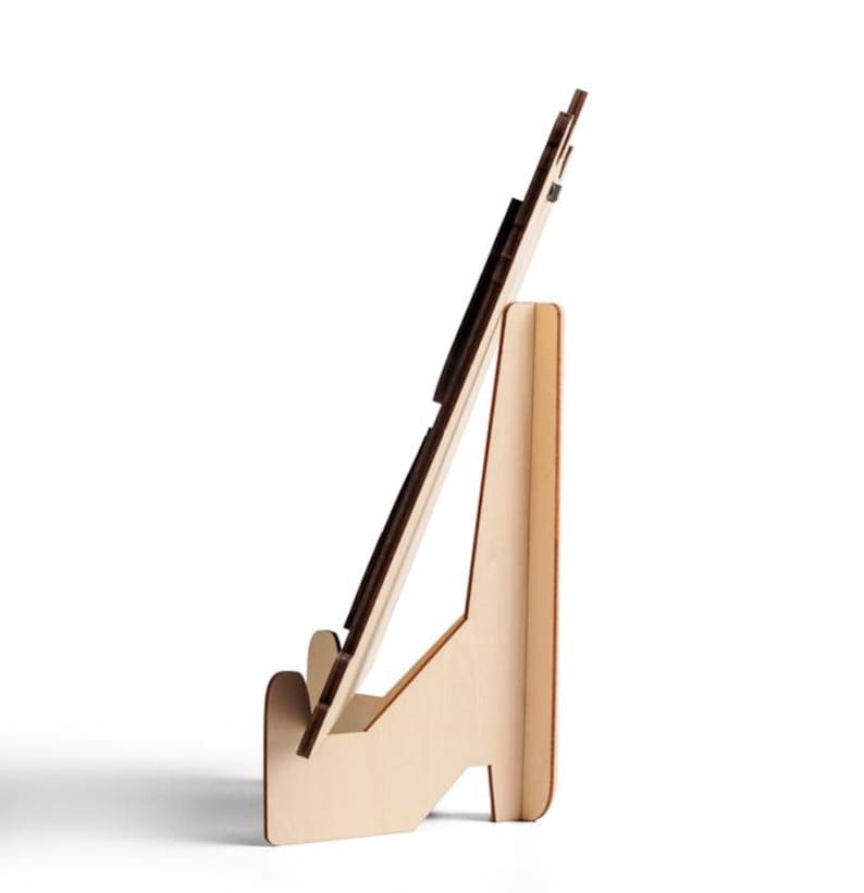 Puede incluir: Un soporte de exhibici&oacute;n de madera de color claro visto desde un perfil lateral. El soporte cuenta con un respaldo vertical y una repisa frontal dise&ntilde;ada para sostener un objeto plano en un &aacute;ngulo inclinado. La construcci&oacute;n consiste en paneles de madera entrelazados.