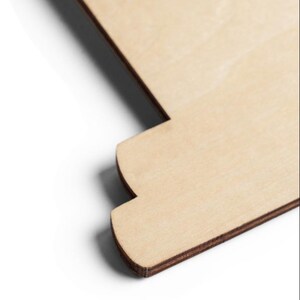 Puede incluir: Un recorte de madera plano y de color claro con una superficie de grano natural y suave. El objeto presenta un perfil de borde escalonado distintivo con una secci&oacute;n de esquina curva, mostrando la textura estratificada del contrachapado en los bordes oscuros.