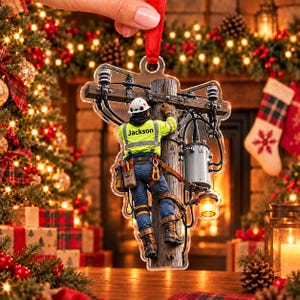 Puede incluir: Un adorno navideño de acrílico transparente que muestra a un operario de líneas eléctricas con una chaqueta amarilla de alta visibilidad con el nombre Jackson impreso en la espalda, jeans azules y un cinturón de herramientas, subiendo un poste.