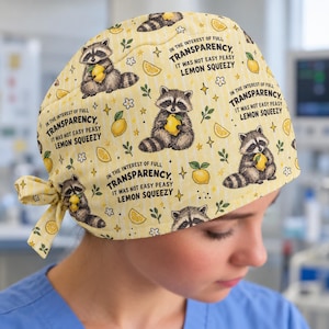 May include: A yellow scrub cap featuring a repeating pattern of raccoons holding lemons, small white flowers, and green leaves. The text "IN THE INTEREST OF FULL TRANSPARENCY, IT WAS NOT EASY PEASY LEMON SQUEEZY" is printed in black across the fabric.