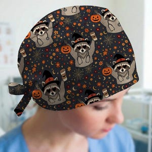May include: A dark gray scrub cap featuring a repeating pattern of raccoons wearing witch hats and heart-shaped glasses. The raccoons hold orange jack-o'-lantern buckets and coffee cups, surrounded by spiderwebs, small orange flowers, and moons.