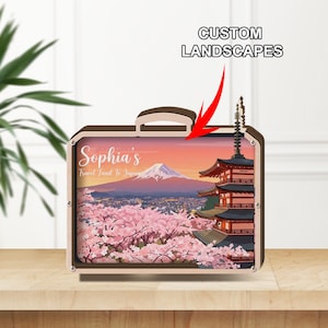 Può includere: Un oggetto decorativo a forma di valigia in legno con un paesaggio giapponese con il Monte Fuji, fiori di ciliegio e una pagoda. Il testo "Sophia's Travel Fund To Japan" è visibile. Sono visibili anche le parole "CUSTOM LANDSCAPES".