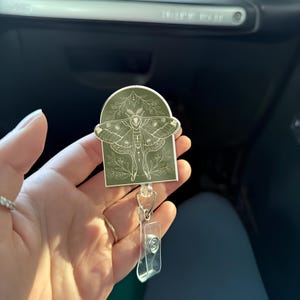 Può includere: Un portabadge retrattile verde e bianco con una grafica di una falena con accenti floreali.
