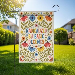 Op de afbeelding: Een tuinvaandel met de tekst "RADICALIZED BY BASIC DECENCY" in kleurrijke letters, omringd door een bloemenontwerp van rode, blauwe, gele en oranje bloemen met groene bladeren. De vlag hangt aan een zwarte metalen standaard in een tuin.
