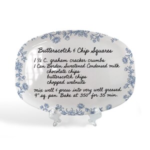 May include: White oval serving platter with a blue floral border and a recipe for Butterscotch & Chip Squares. The recipe includes ingredients like graham cracker crumbs, chocolate chips, and walnuts. The platter is displayed on a clear stand.