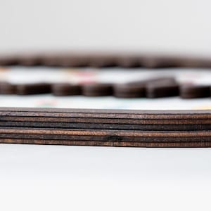 Puede incluir: Un marco de madera marr&oacute;n oscuro con una construcci&oacute;n de bordes superpuestos. El marco presenta un acabado liso y pulido con capas de veta de madera visibles en el lateral. Una superficie blanca con patrones coloridos borrosos es visible dentro.
