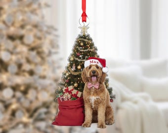 May include: A Christmas ornament featuring a golden doodle dog wearing a Santa hat with the name "Max" and a red and white striped bow tie. A red sack of gifts and a decorated Christmas tree are in the background.