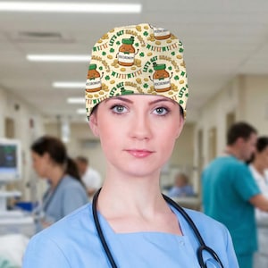 Może przedstawiać: Medyczny czepek chirurgiczny z powtarzającym się wzorem z kreskówkowych słoików, koniczyny i złotych monet na kremowym tle. Słoiki mają napis "Rocuronium" i frazę "Let's Get Sham-Roc'd". Czepek jest noszony przez osobę w niebieskim topie.