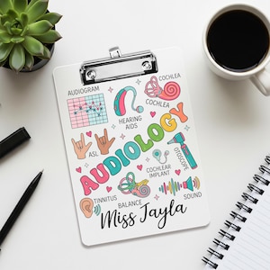 Può includere: Un appunti bianco con una clip argentata, con illustrazioni colorate e la parola "AUDIOLOGY" a lettere grandi. L'appunti include grafica medica, testo e il nome "Miss Jayla". Sono visibili anche una tazza di caffè e una pianta succulenta.