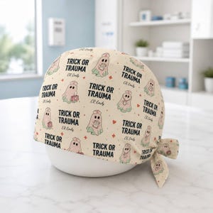 May include: A cream-colored scrub cap featuring a repeating pattern of ghosts dressed as nurses. Each ghost holds a medical clipboard or kit. The text reads TRICK OR TRAUMA and ER Emily in black font, accented by small red medical crosses and hearts.