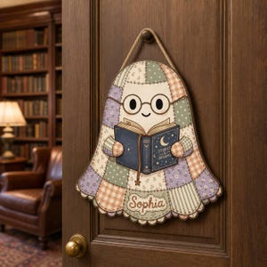 May include: A bell-shaped wall hanging featuring a ghost character with round glasses, reading a dark blue book titled Stories & Dreams. The ghost has a patchwork pattern in muted pastel colors and the name Sophia printed at the bottom edge.
