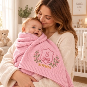 Könnte beinhalten: Eine weiche, gestrickte Babydecke in Rosa mit einem gestickten Wappen, das den Buchstaben S, eine Schleife und florale Akzente zeigt. Der Name SOPHIA ist in Goldfaden unter dem Wappen eingestickt. Die Decke hat einen gerippten Rand und ein Strickmuster.