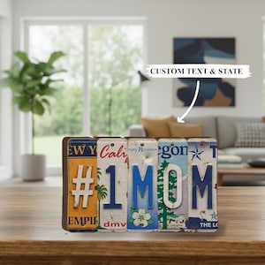 May include: A decorative sign spelling "#1 MOM" using repurposed license plates. The sign features plates from New York, California, Michigan, Oregon, and Texas. The sign is displayed on a wooden table.