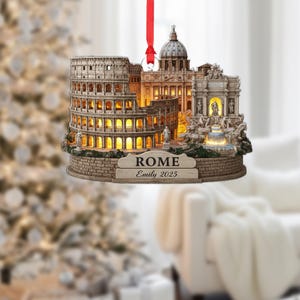 May include: A Rome-themed Christmas ornament depicting the Colosseum, St. Peter's Basilica, and the Trevi Fountain. The ornament has a warm, illuminated glow and includes the name "Rome" and the name "Emily 2025". It hangs from a red ribbon.