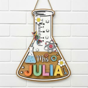 May include: Wooden science-themed wall hanging in the shape of a flask. Features a white background with colorful accents, including a DNA strand, atom, and the name "JULIA" in bright letters. Includes the text "Mrs."