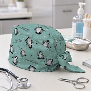 May include: A teal scrub cap featuring a repeating pattern of cartoon penguins holding knives, interspersed with small hearts and stars. The text "flight was never an option" is printed in black script throughout the fabric design.