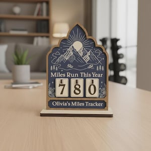 Peut inclure: Compteur de miles en bois avec un fond bleu marine et un motif de montagne et de soleil. Le texte "Miles Run This Year" et "Olivia's Miles Tracker" sont visibles. Le compteur affiche "780" miles.