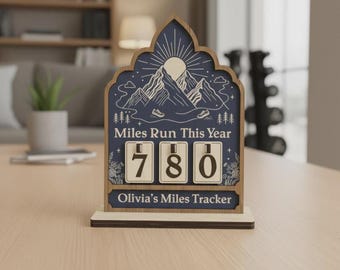 May include: Wooden miles tracker with a navy blue background and a mountain and sun design. The text "Miles Run This Year" and "Olivia's Miles Tracker" are visible. The counter shows "780" miles.