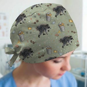 May include: A sage green scrub cap featuring a repeating pattern of raccoons, silver trash cans, gold crowns, brown sunglasses, banana peels, and small paw prints. The fabric has a soft, matte finish with a tie-back closure at the nape of the neck.