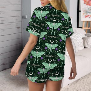 May include: A short-sleeved pajama set featuring a dark green background with a repeating pattern of light green luna moths, purple flowers, white crescent moons, and the name Sophia printed in a white cursive script throughout the fabric.