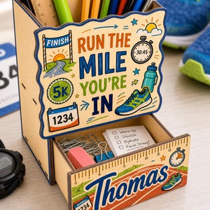 Puede incluir: Organizador de escritorio de madera con temática de running. El organizador presenta el texto "Run the Mile You're In" e incluye un cajón con útiles de papelería. El diseño incorpora una zapatilla de running, un cronómetro y un gráfico de línea de meta. También se muestra el nombre "Thomas".