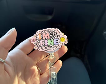 May include: A pink retractable badge holder with a black and white doodle design. The holder has the text "I'M FINE" in colorful letters.