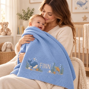 Puede incluir: Una manta de punto para bebé en color azul claro con un bordado de temática oceánica que incluye una ballena, una tortuga marina, conchas y estrellas de mar. El nombre FINN está bordado en el centro con hilo claro, rodeado de plantas acuáticas.