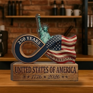 May include: Wooden commemorative sculpture featuring the Statue of Liberty, an American flag, and the text "250 YEARS OF FREEDOM." The base reads "UNITED STATES OF AMERICA" with the years "1776 2026." The sculpture is made of wood and has a rustic appearance.