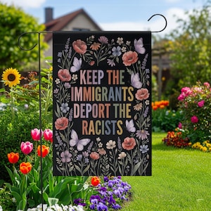 May include: Black garden flag with the text "KEEP THE IMMIGRANTS DEPORT THE RACISTS" surrounded by colorful flowers and butterflies. The flag is displayed in a garden with flowers and greenery.