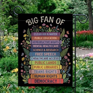 May include: Black garden flag with the text "BIG FAN OF" above a stack of colorful books. Each book lists a different cause, including "CLEAN AIR & WATER" and "DEMOCRACY". The flag is decorated with flowers and butterflies.